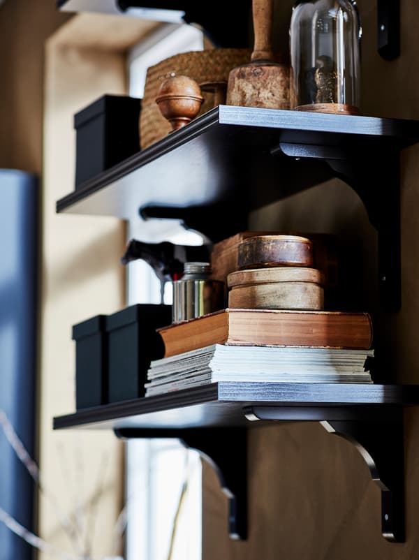 Estante de parede BERGSHULT/RAMSHULT escura com uma pilha de revistas e livros, caixas e arrumação para exposição.