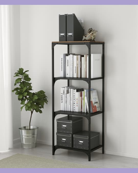 Estante vertical de metal negro con cuatro niveles, organizado con libros, archivadores grises y cajas. A un costado hay una planta en macetero metálico.