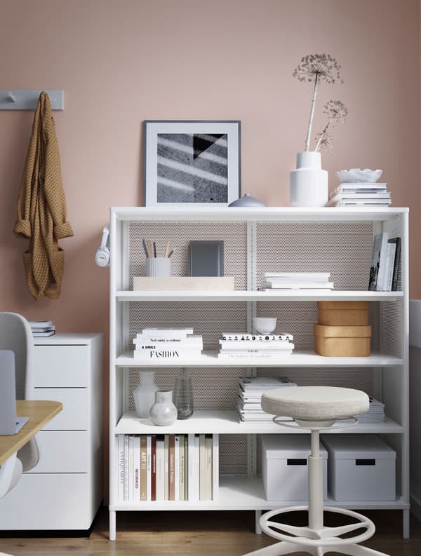 Estantería BEKANT blanca con libros, jarrones, material de oficina, auriculares y cajas de almacenaje. Hay un taburete de color crema delante.