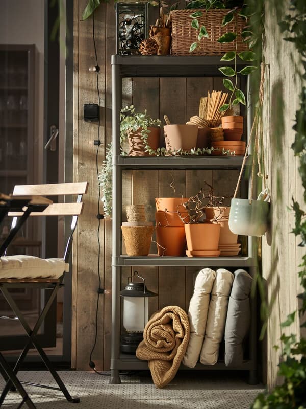 Estantería exterior con cojines, plantas, maceteros, lámpara, manta, cesta de mimbre y elementos de decoración. 