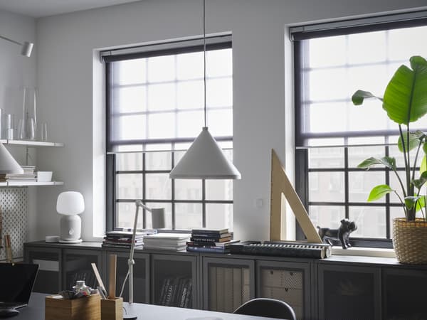 Estores de correr transparentes que cobrem metade de duas janelas, armários em cinzento com portas em rede, um candeeiro suspenso branco e uma planta verde.