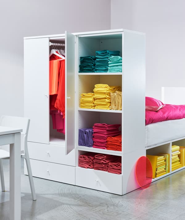 Estrutura de cama com um roupeiro integrado a servir de cabeceira de cama com roupa colorida arrumada no interior.