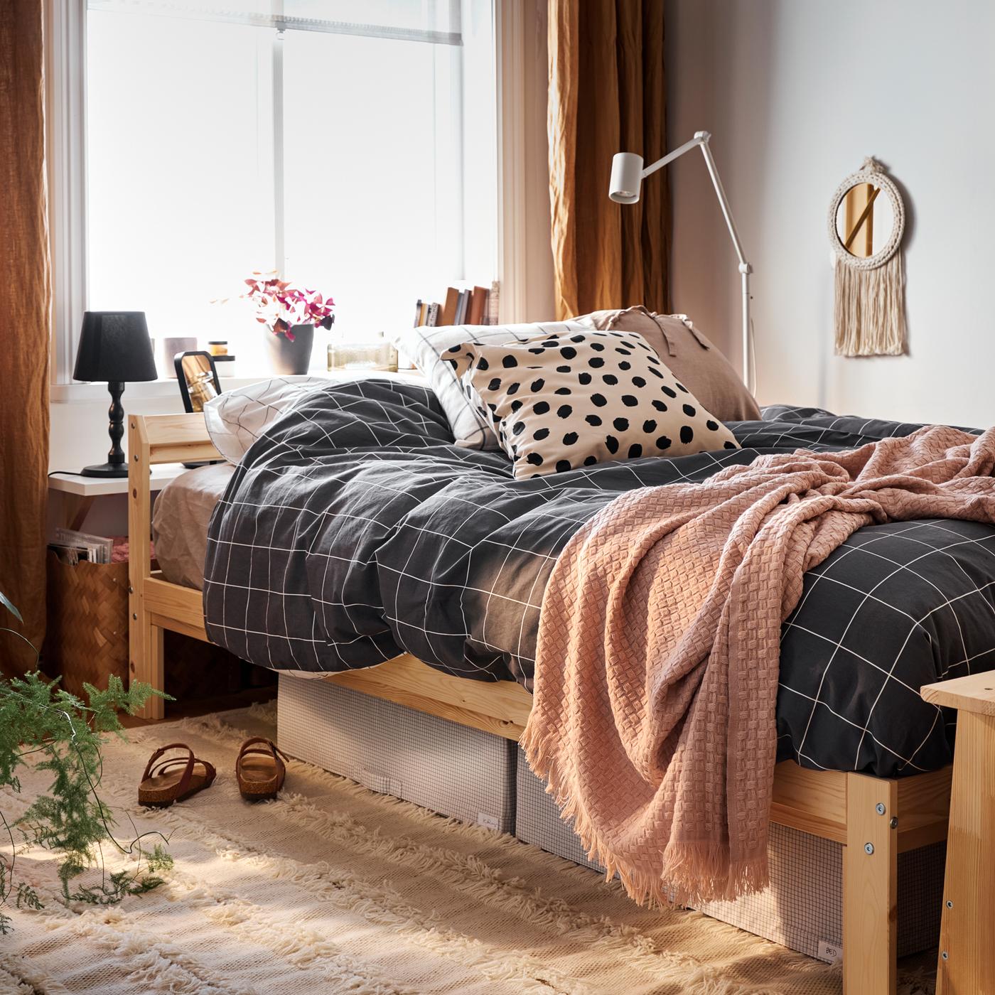 Estrutura de cama de piñeiro con roupa de cama a cadros en branco e negro, con caixas de almacenaxe en branco/gris e unha alfombra debaixo.