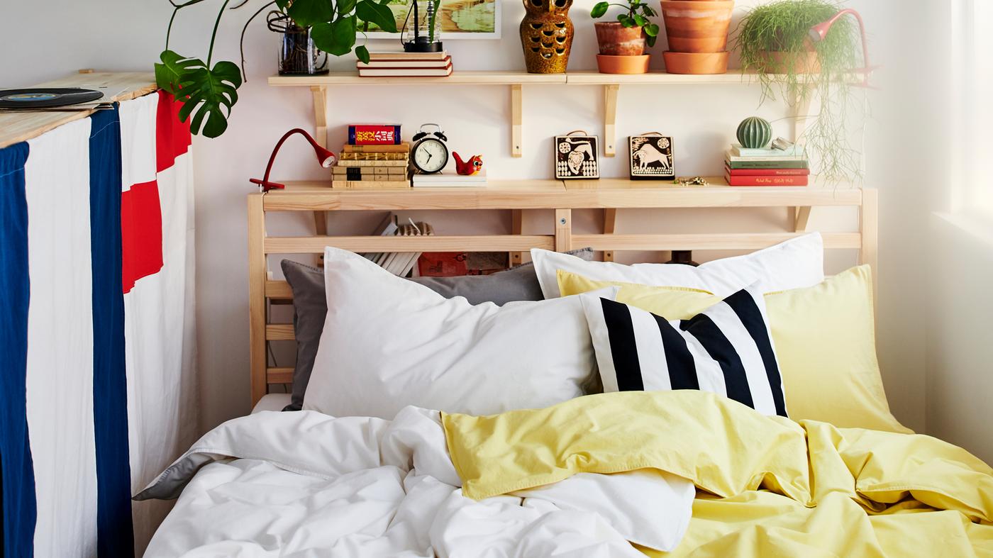 Estrutura de cama TARVA em pinho, com roupa de cama ÄNGSLILJA em branco e amarelo claro, encostada a parede branca com prateleiras em choupo.