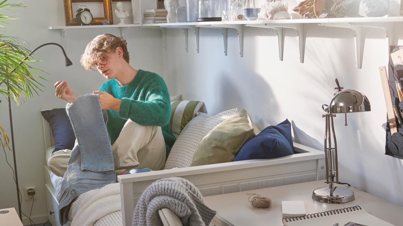 Estudante sentado numa cama individual/dupla HEMNES a coser um par de calças de ganga. Prateleiras na parede com um relógio despertador, molduras e objetos.