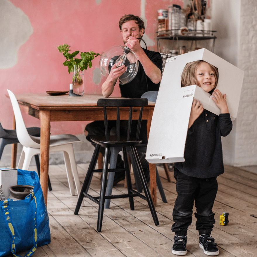 Et barn har taget en IKEA papkasse over hovedet, mens han far samler en lampe ved spisebordet i baggrunden.