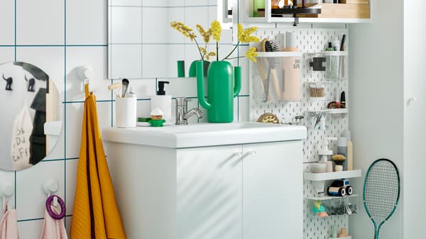 Et hvidt ENHET skab til vask med to låger, gule og lyserøde håndklæder, en grøn vase og en SKÅDIS hulplade med toiletartikler.