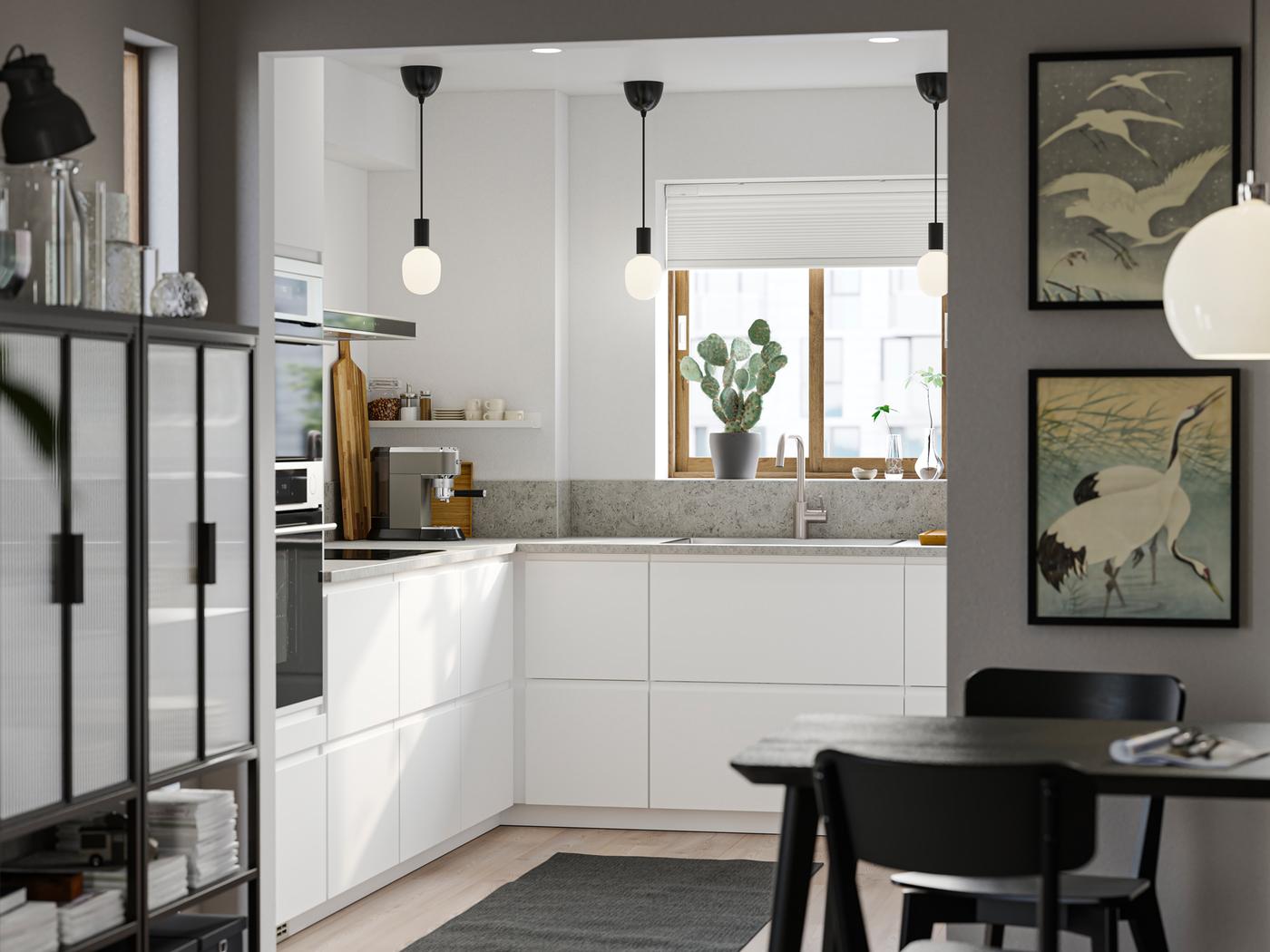 Et lille køkken med strømlinede, matte hvide VOXTORP fronter har tre sorte loftslamper og et spiseområde i forgrunden.