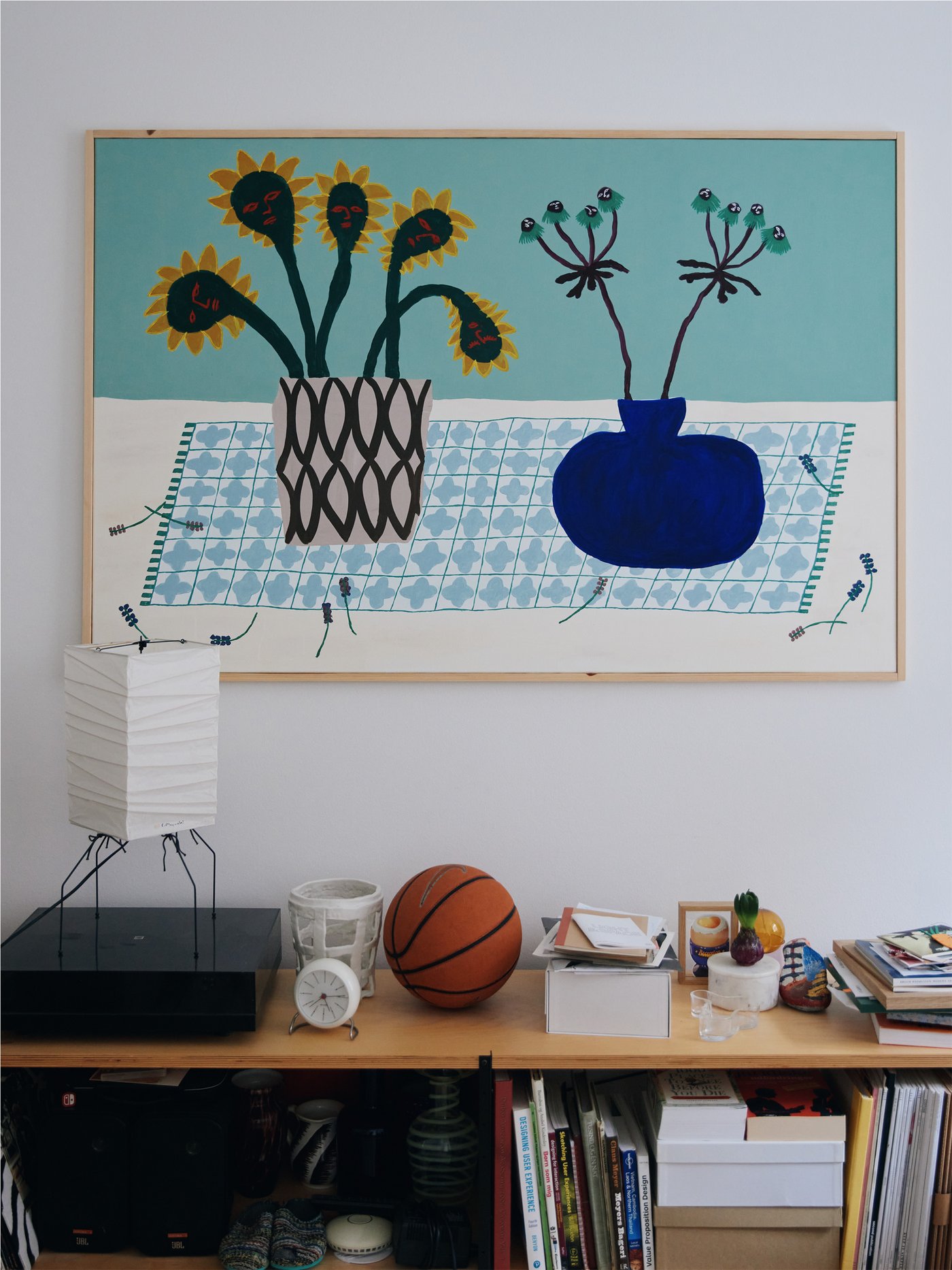 Et maleri med en turkis bagrund og en blå vase i en træramme er i fokus og billedet hænger over en reol fyldt med ting.