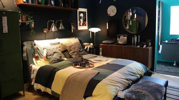 Et mørk og intimt soveværelse med sorte vægge og en seng i fokus og på sengen står en bakke over sengen hænger to lamper og til højre for sengen står en kommode og et STOCKHOLM spejl.