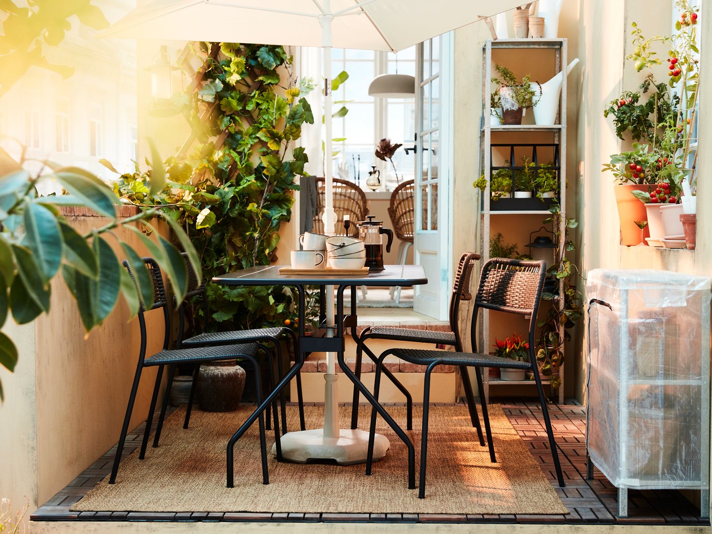 Et solbeskinnet uderum med et mørkegråt VIHOLMEN havemøbelsæt, en TVETÖ parasol, masser af planter og et LYDERSHOLM tæppe.