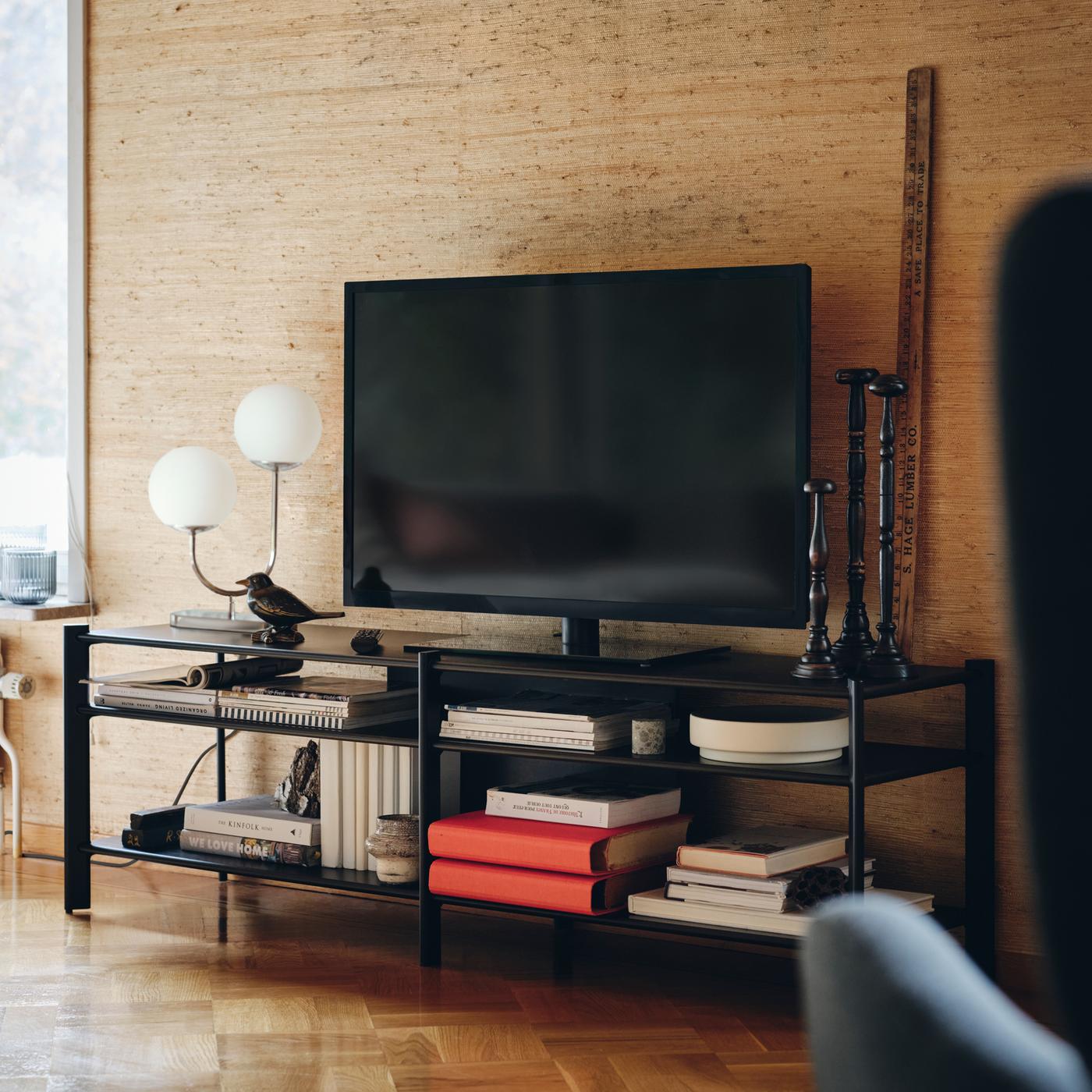 Et sort tv står mellem en bordlampe og tre lysestager på et sort JÄTTESTA tv-bord på et trægulv.