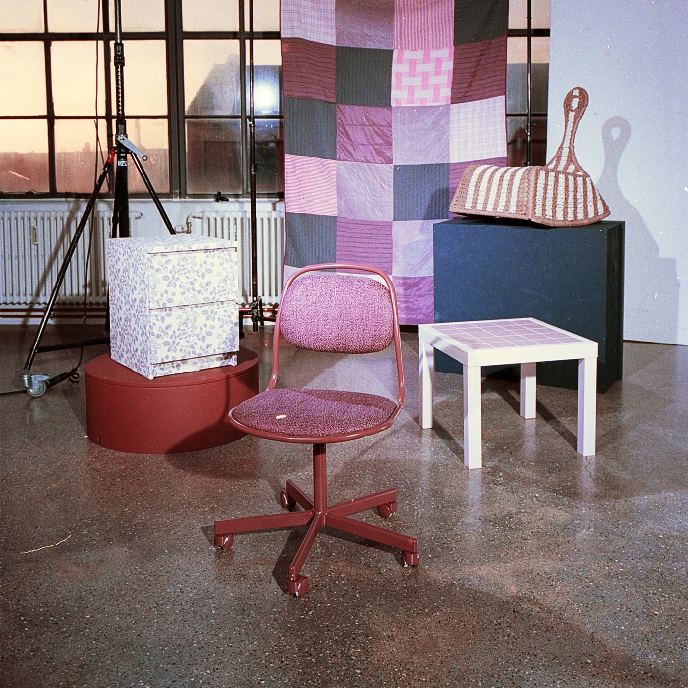 Et tæppe hænger i baggrunden mens der foran står en hvid kommode en pink kontorstol et hvidt sidebord og en hvidstribet fletstol på et blåt podie som alle er del af FÖRNYA projektet hos IKEA.