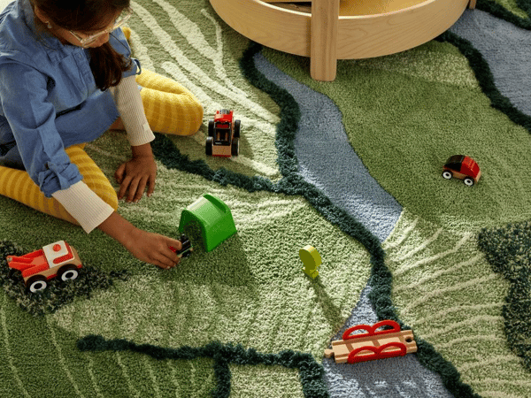 Ett barn leker med röda tågleksaker i trä på en matta som ser ut som en karta över land och sjöar, grönt och blått.