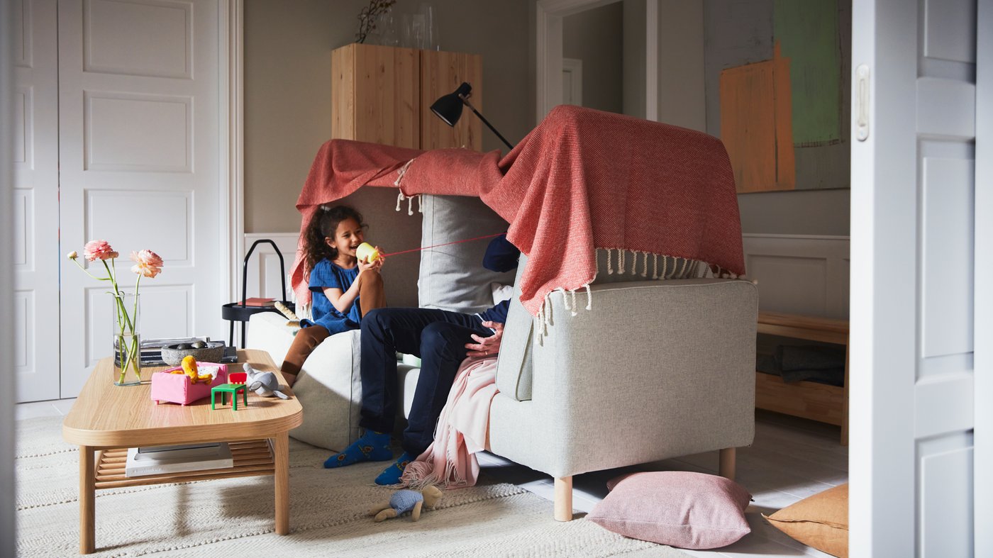 Ett barn och en vuxen pratar i en trådtelefon i ett tält gjort av en beige PÄRUP soffa och en rödbrun/vit ULLTISTEL pläd.