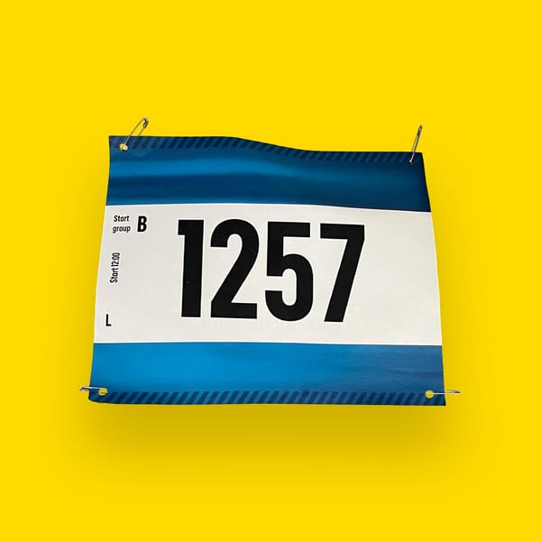  Ett blått och vitt startnummer med siffran ”1257” och texten ”Start group B” samt ”L”, fäst med säkerhetsnålar, mot gul bakgrund.
