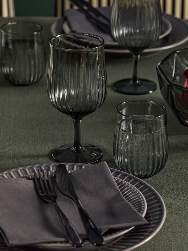 Ett bord är dukat med mörkgrå linneduk och mörkgrå porslin med svarta bestick som ligger på linneservetter. vinglas och selterglas i mörkgrått räfflat glas med vatten i står på bordet.