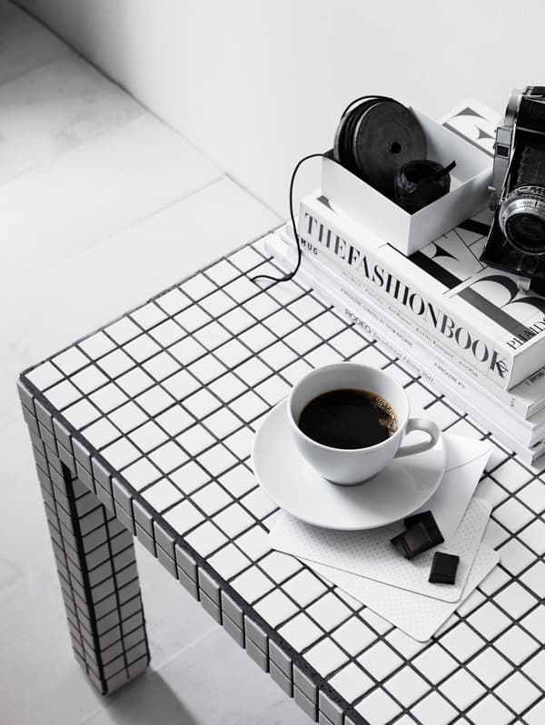Ett bord med svartvitt rutmönster. På bordet står en vit kopp med svart kaffe på ett vitt fat. Bredvid ligger två små chokladbitar på en vit servett. I bakgrunden till höger syns en stapel med böcker.