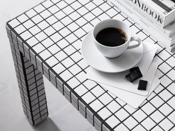 Ett bord med svartvitt rutmönster. På bordet står en vit kopp med svart kaffe på ett vitt fat. Bredvid ligger två små chokladbitar på en vit servett. I bakgrunden till höger syns en stapel med böcker.