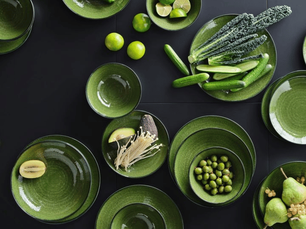 Ett buteljgrönt porslin med skålar, fat och assietter står på ett bord med en svart duk. På några av skålarna och faten ligger det citroner, oliver, små gurkor eller päron på.