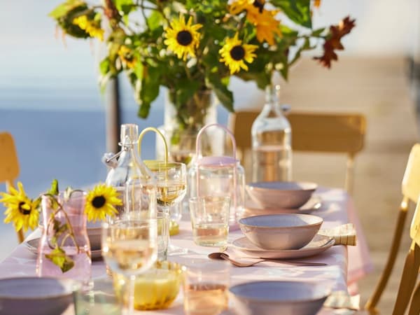 Ett dukat bord med sommarblommor och solrosor i en vas, omgiven av tallrikar, glas och karaffer under blå himmel.