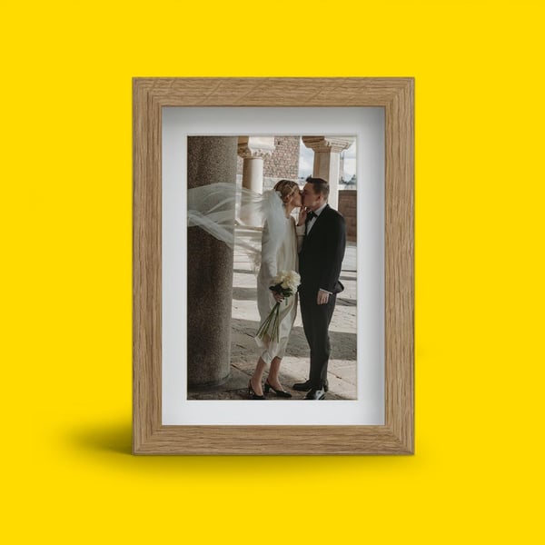 Ett foto i en träram som visar ett brudpar stående nära varandra, med brudens slöja som blåser i vinden, mot gul bakgrund.