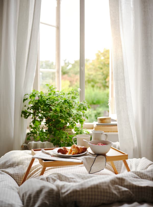 Ett frukostbord för sängen står på ett fluffigt vitt täcke på en säng, framför ett öppet fönster
