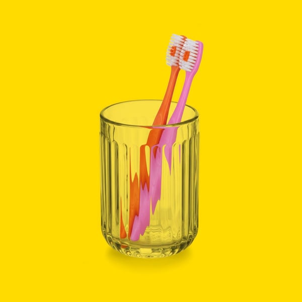 Ett glas med två tandborstar, en rosa och en orange, mot gul bakgrund.