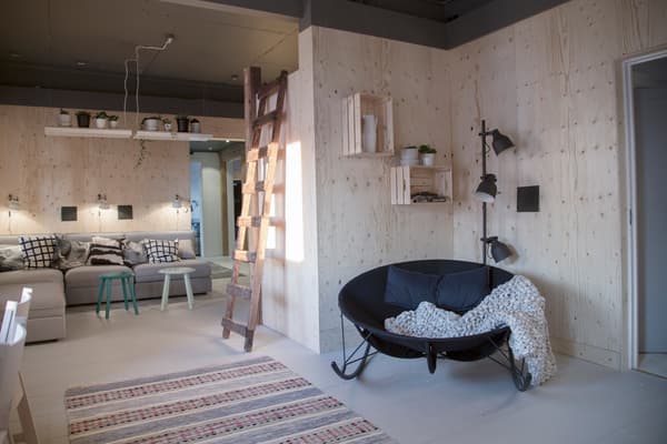 Ett modernt och hemtrevligt vardagsrum med träväggar och ljust golv. En svart rund fåtölj med två mörkblå kuddar och en vit filt står i förgrunden. En trästege leder upp till ett loft, och intill finns vägghyllor. I bakgrunden syns en grå soffa med mönstrade kuddar och två små bord framför. Ovanför soffan hänger enkla lampor.