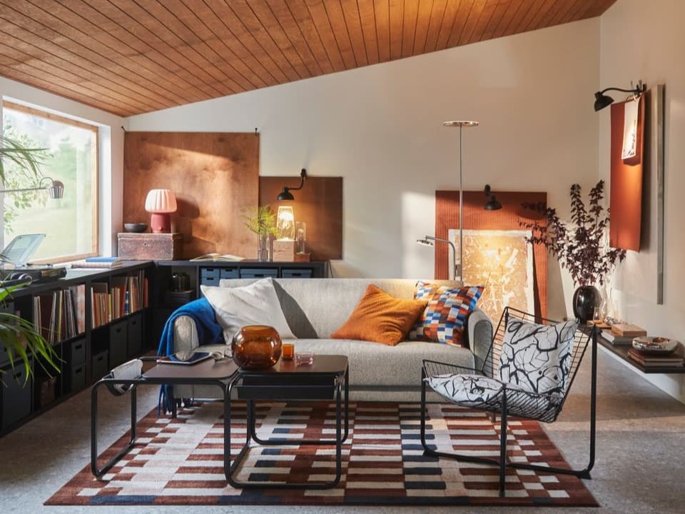 .Ett modernt vardagsrum med en grå soffa, ett mönstrat soffbord, och en dekorativ matta. Det finns växter i krukor och tavlor lutade mot väggen.