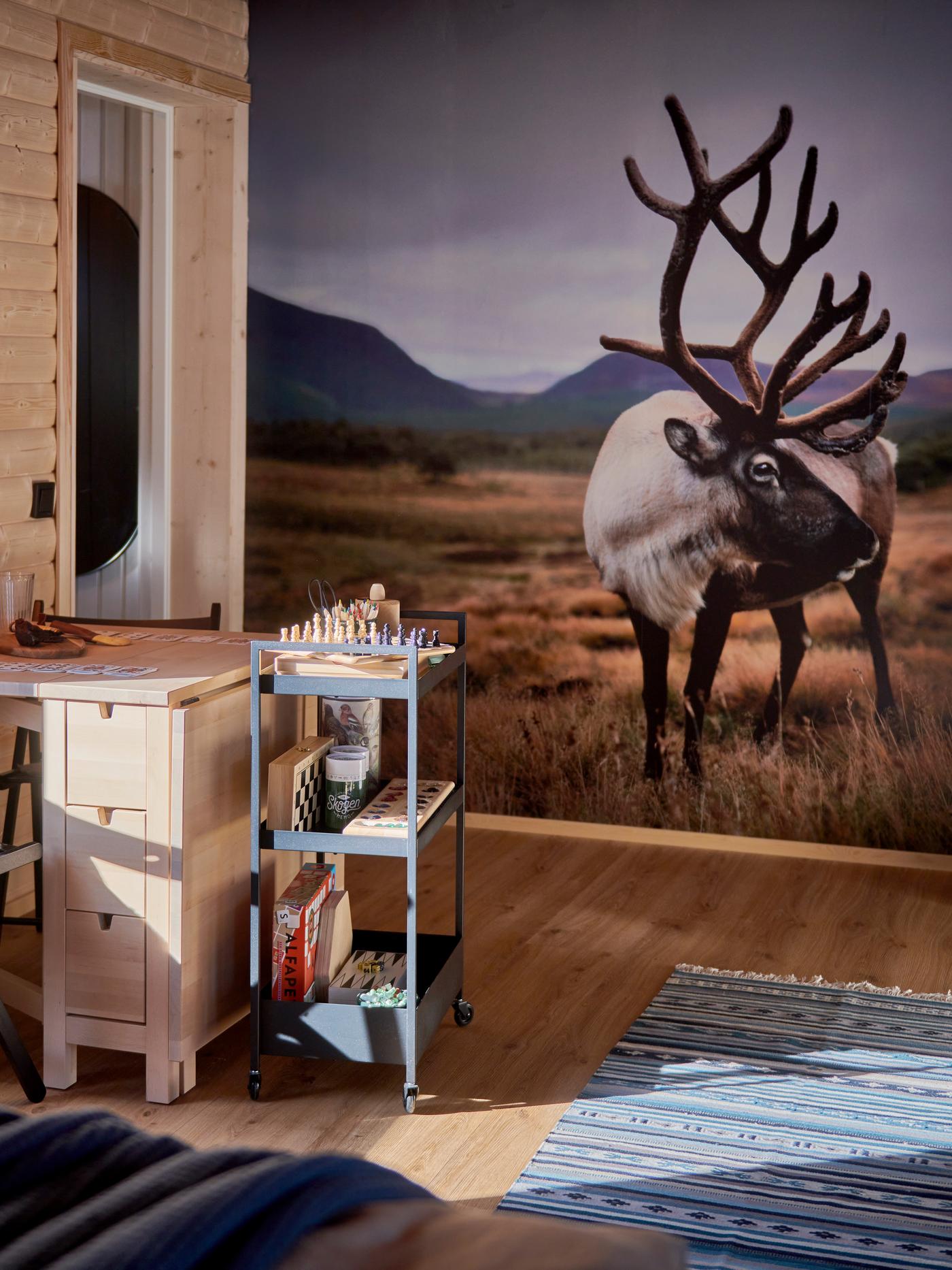 Ett ombonat rum med träväggar, ett skrivbord med ett schackspel, en metallvagn med böcker och en stor väggmålning av en ren i fjällandskap. En randig matta syns i förgrunden.