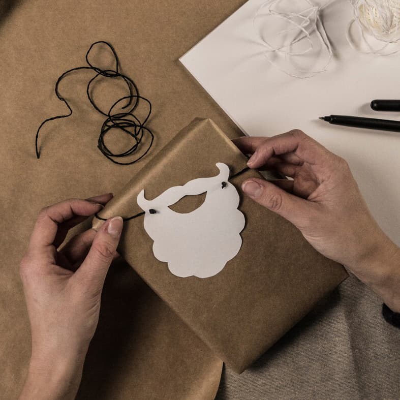 Ett par händer knyter fast ett tomteskägg utklippt ur ett vanligt papper på ett brunt paket.