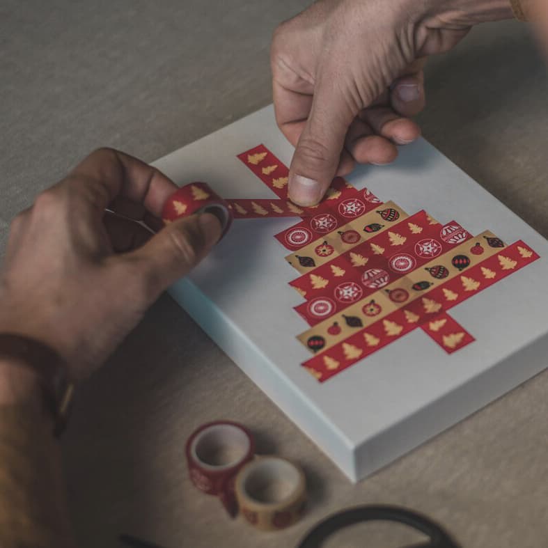 Ett par händer tejpar formen av en gran med washitejp på en vit papplåda.