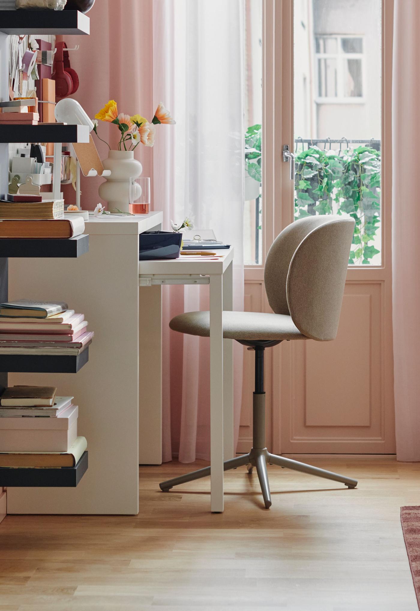 Ett stilrent hemmakontor med ett vitt LÄNGDHOPP skrivbord, en modern beige snurrstol och svarta hyllor, mot en rosa vägg och en glasdörr som leder ut till en balkong med gröna växter.