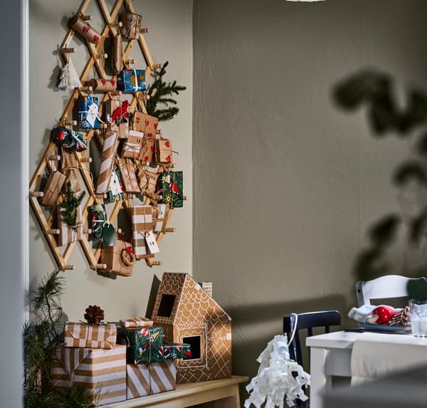Ett väggmonterat julträd av trä med små presenter, bredvid ett bord och en stapel med julklappar.