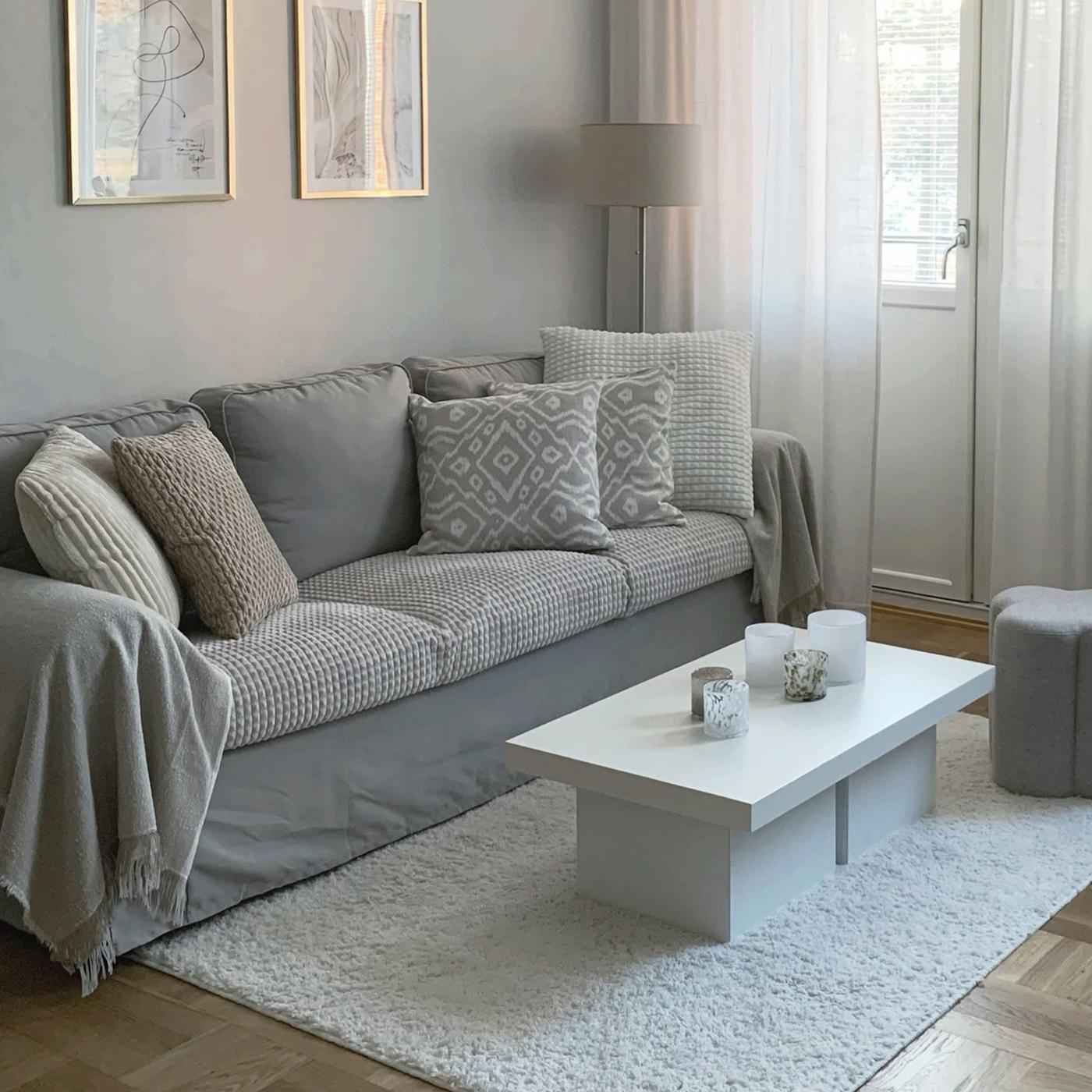 Ett vardagsrum med en grå 3-sits soffa, vitt soffbord och ljusa gardiner. Soffan är dekorerad med kuddar och filtar i färgerna grått, vitt och beige. 