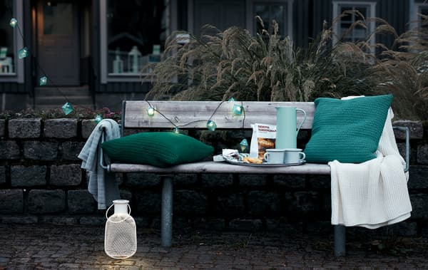 Extérieur, décor de début de soirée en ville avec un banc public aménagé avec un plateau à café, des coussins, un plaid et des éclairages décoratifs.
