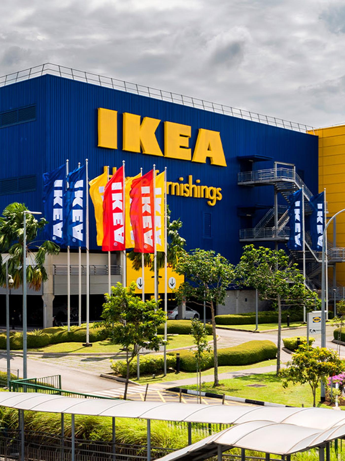 Exterior of IKEA Tampines, with "IKEA" logo on the blue walls. There are red, blue and yellow flags with white "IKEA" logo raised in front of the building.
