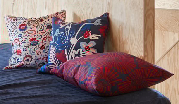 Extremidade ou lateral de uma cama arrumada, junto a uma parede em contraplacado. Três almofadas com padrões elaborados a decorar a cama.