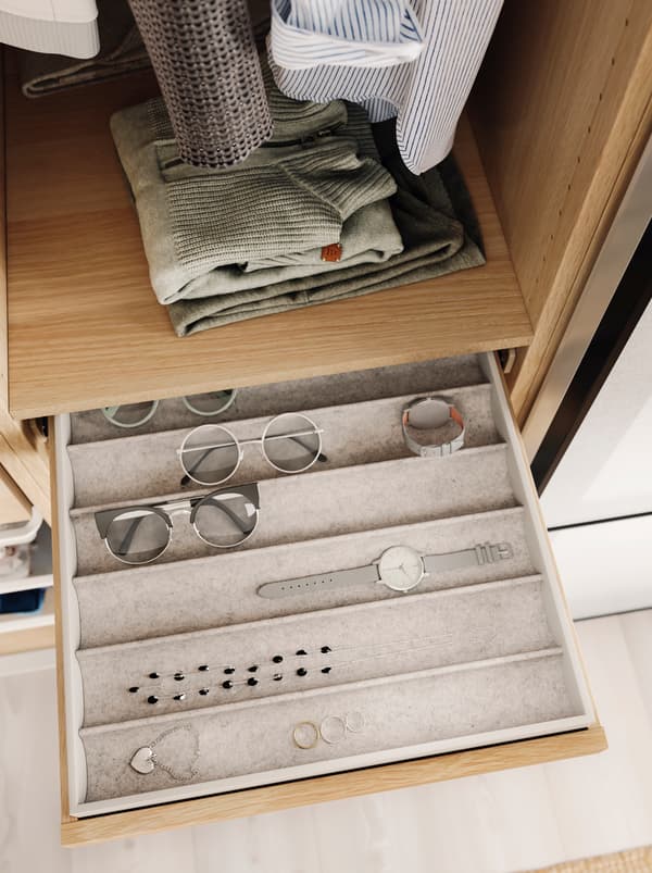 Eyeglasses, a watch, and jewellery in neat rows in a KOMPLEMENT insert within a pulled-out, shallow drawer in an open wardrobe.