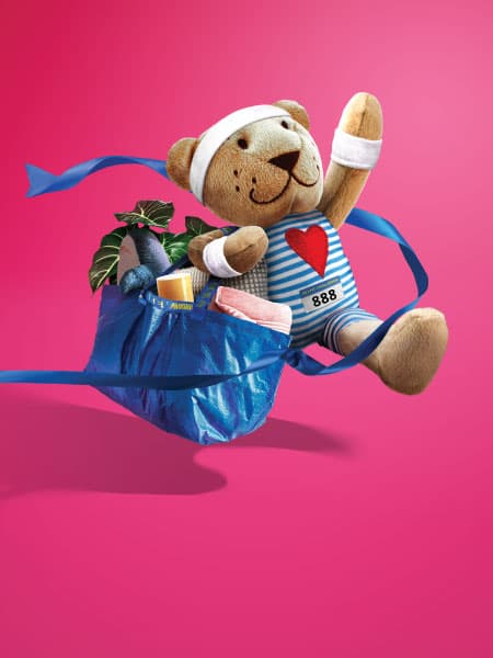 FABLER BJÖRN teddy bear soft toy wearing a white headband and wristbands, running with a blue FRAKTA bag filled with household items on a pink background.