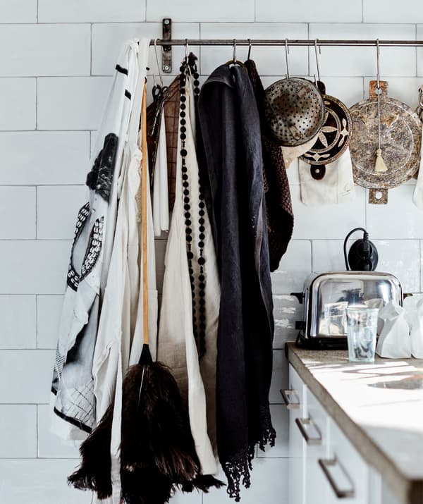 Fabric, a duster and colanders hanging on a metal rail on a white tiled wall.