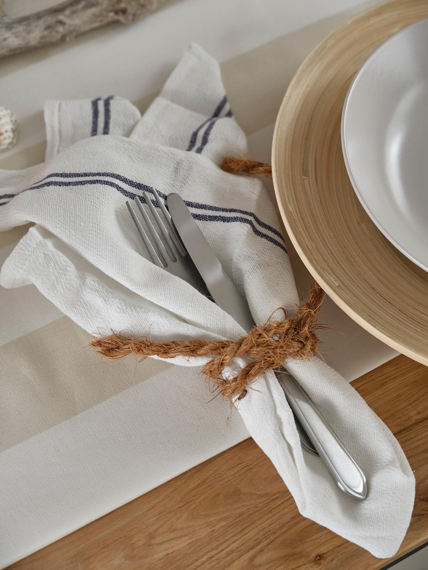Fabric napkin with cutlery and jute tie beside a white plate on a wooden table setting.