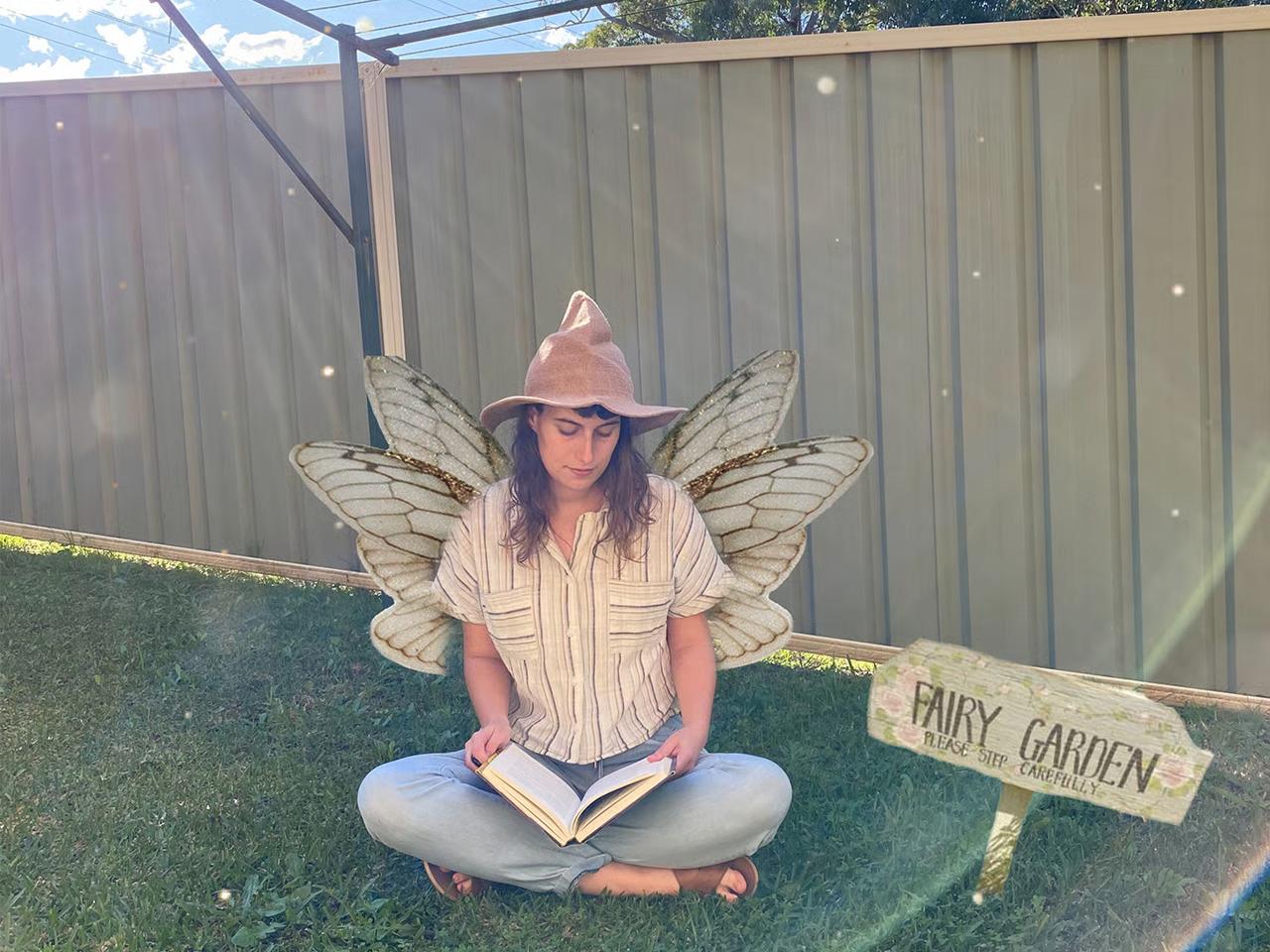 אישה יושבת על רצפה במרפסת שמכוסה בדשא מלאכותי. היא קוראת ספר. יש עליה כובע קוסמים וכנפי פרפר. לידה יש שלט שכתוב עליו בכתב יד Fairy Garden (גן הפיות).