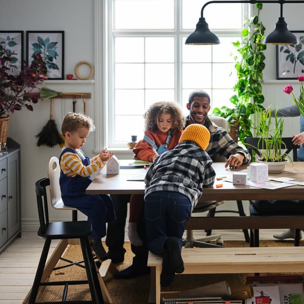 Família de três crianças pequenas e um pai sentados numa mesa de refeição em madeira com uma janela e vasos com plantas em segundo plano.