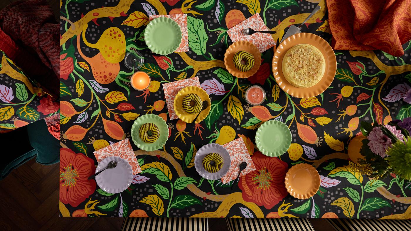 Farbenfroher IKEA Esstisch mit lebendig gemusterter Tischdecke in floralen Motiven, gedeckt mit buntem Geschirr, Papier-Servietten und einem Kuchen – ein einladendes Tischarrangement von oben fotografiert.