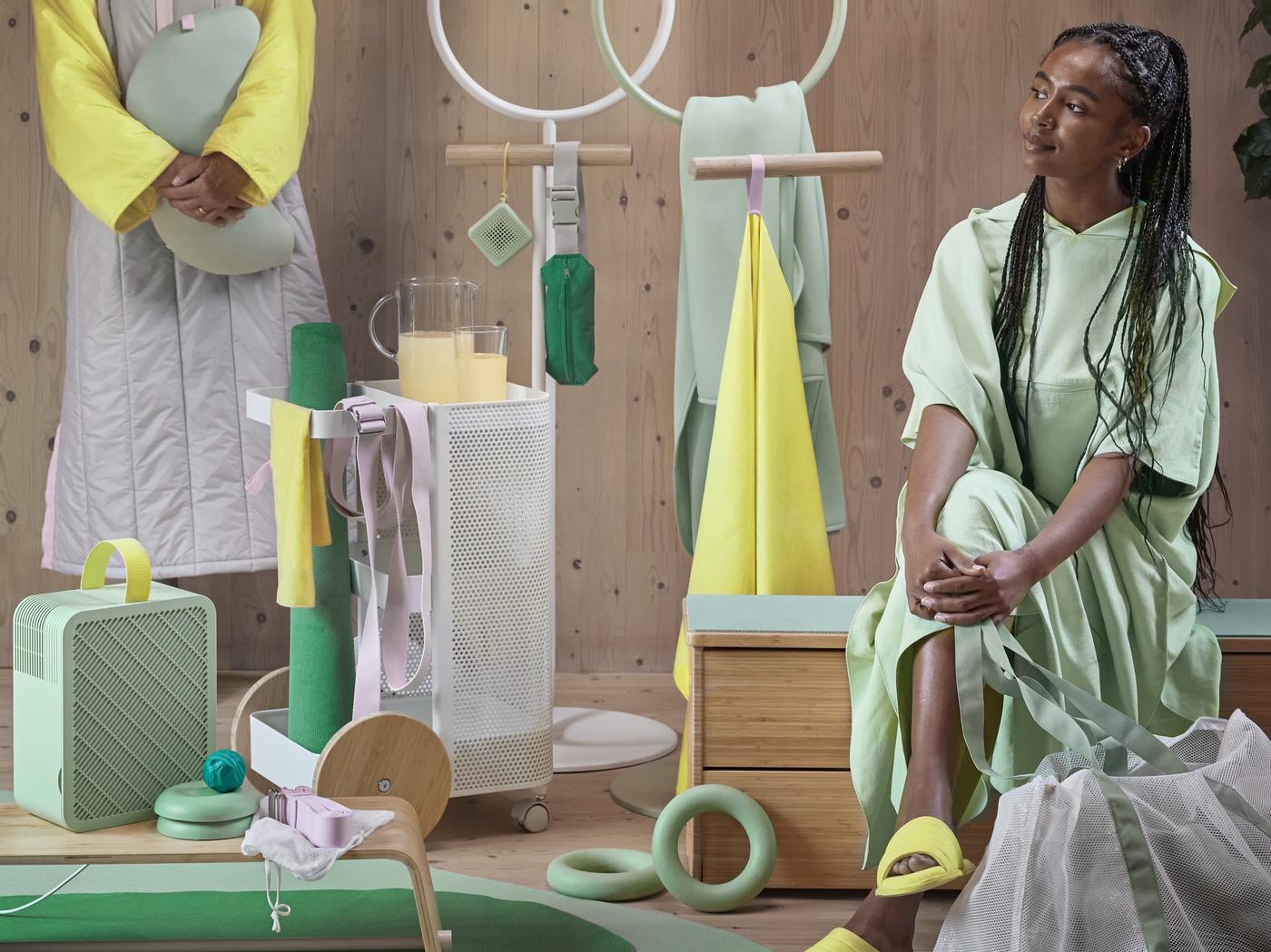 Farblich abgestimmte IKEA Szene mit modernen Wohnaccessoires in Pastellgrün, Gelb und Beige; zwei stilvoll gekleidete Frauen mit IKEA Textilien, Aufbewahrungselementen, Hockern und einem tragbaren Lautsprecher; betont wird kreatives, farbenfrohes Einrichten mit IKEA.“
