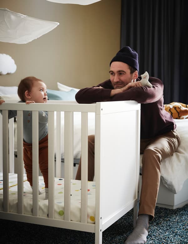 Father looking at son who is standing in an IKEA SUNDVIK cot with spotted bed sheet in a bedroom.