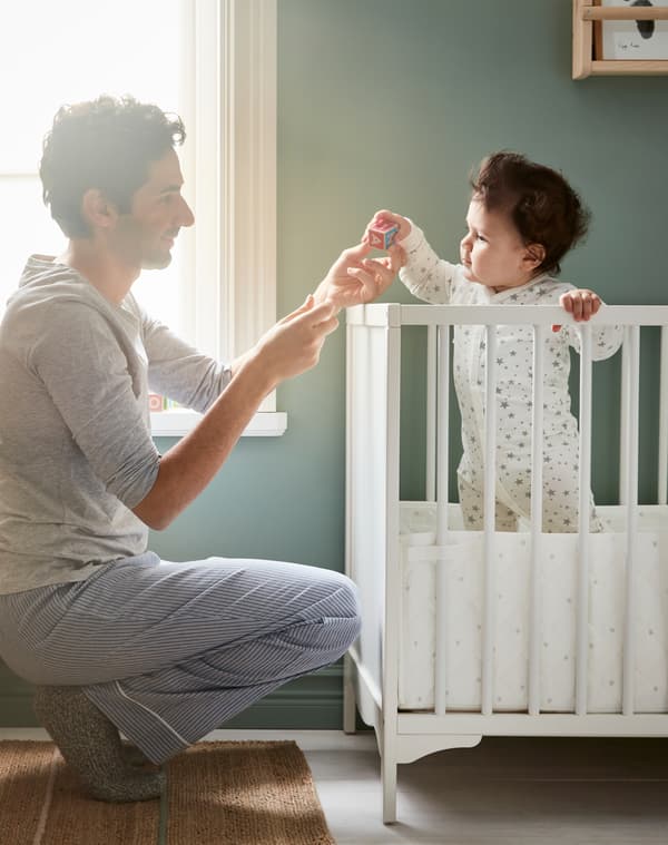 Father playing with a baby and their small soft toy while the baby stands in a white IKEA SUNDVIK cot against window light.
