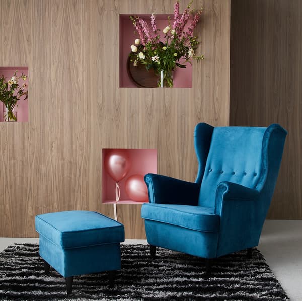 Fauteuil à oreilles STRANDMON en velours bleu avec repose-pieds, posé sur un tapis noir et blanc.